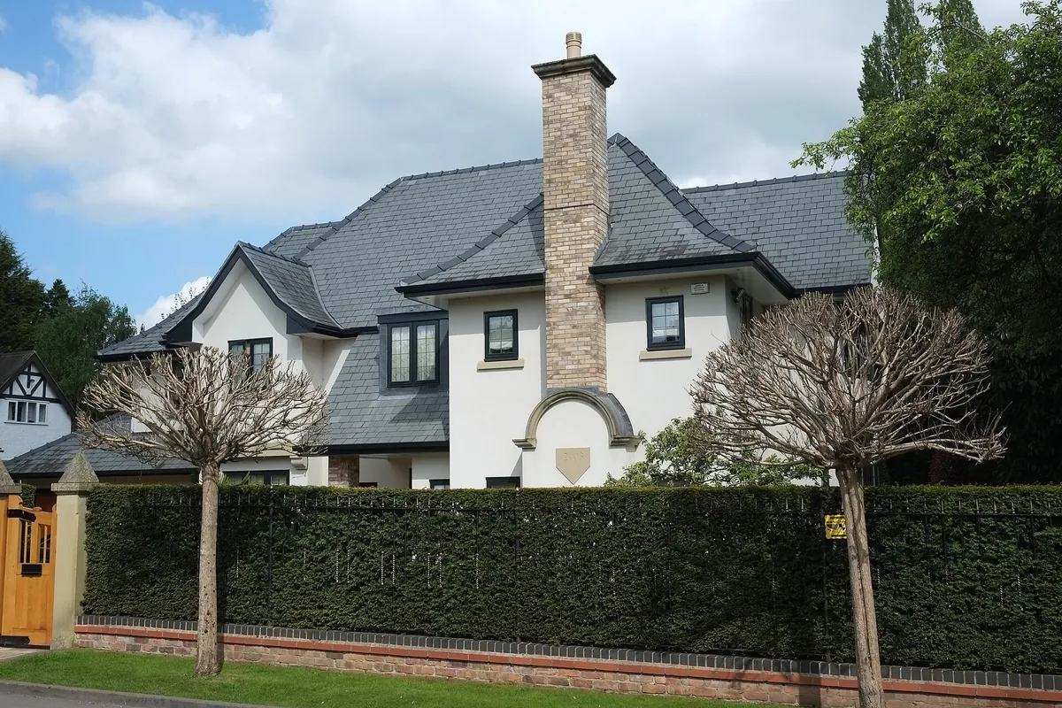 A large white-rendered house with a tall stone chimney behind a clipped hedge in Bowdon